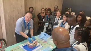 Group of clinicians gathered around a table watching a hands-on demonstration of a peritoneal dialysis catheter insertion technique.