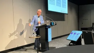 Paul Bennett presenting at a conference with presentation screens displaying information about assisted peritoneal dialysis.
