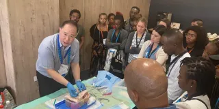Group of clinicians gathered around a table watching a hands-on demonstration of a peritoneal dialysis catheter insertion technique.