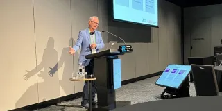 Paul Bennett presenting at a conference with presentation screens displaying information about assisted peritoneal dialysis.