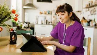 A women wearing scrubs sits in her kitchen taking notes while watching her tablet