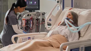 A healthcare professional supports a patient at the bedside with a PrisMax machine behind her
