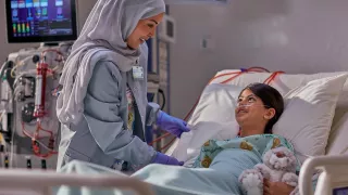 A clinician talks to a young patient who is lying in their hospital bed. There is a PrisMax machine in the background.