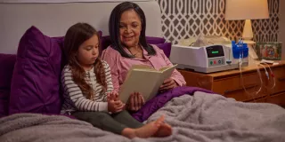 A women is reading to her granddaughter in her bed. The Homechoice Claria system sits on her nightstand