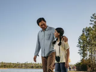A man and a young girl walking by a serene lake, enjoying the peaceful surroundings and each other's company.